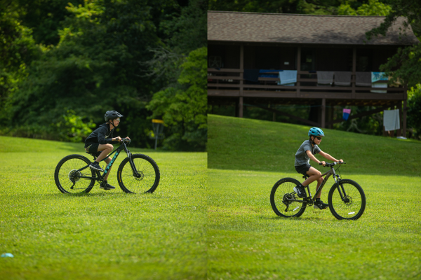 Two children riding bikes on a sunny grassy field; left rider in black gear, right rider in gray shirt with a blue helmet, with a wooden building in the background.