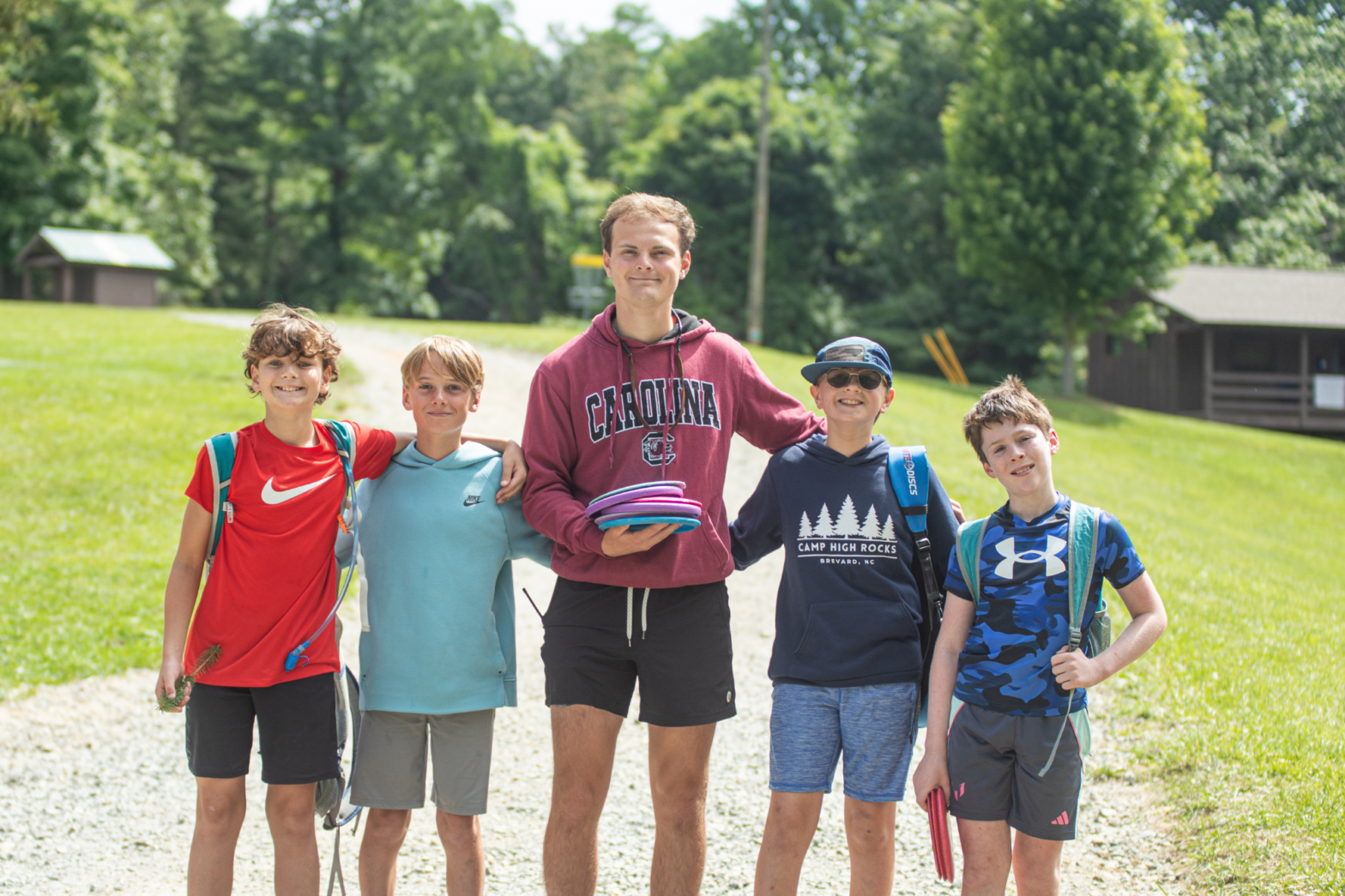 Group of five campers standing arm in arm on a sunny field, smiling at the camera.
