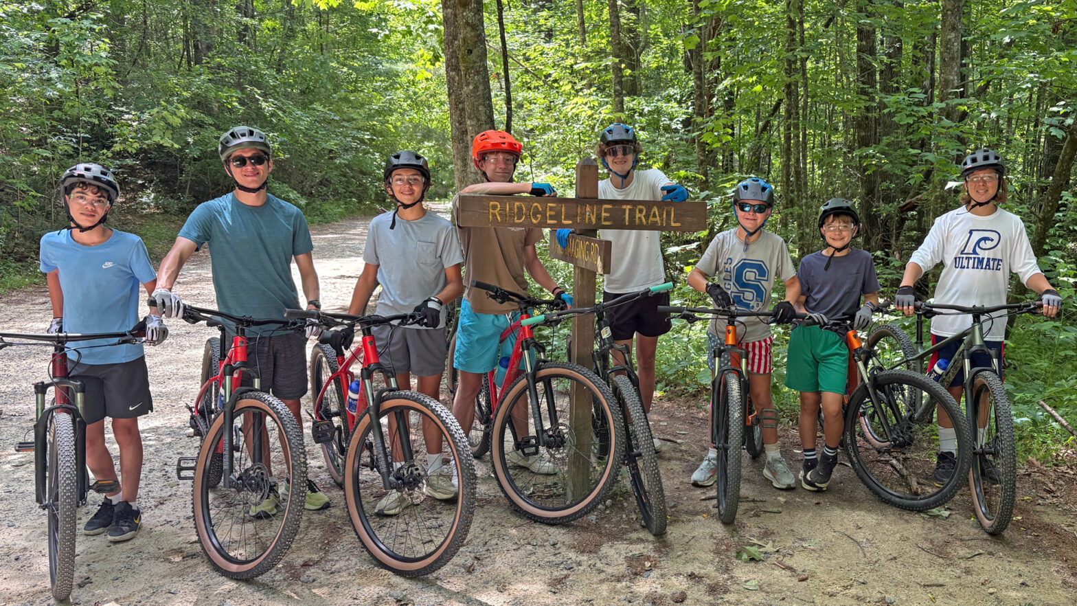Eight children wearing helmets pose with their mountain bikes on a dirt forest trail beside a Ridgeline Trail sign.