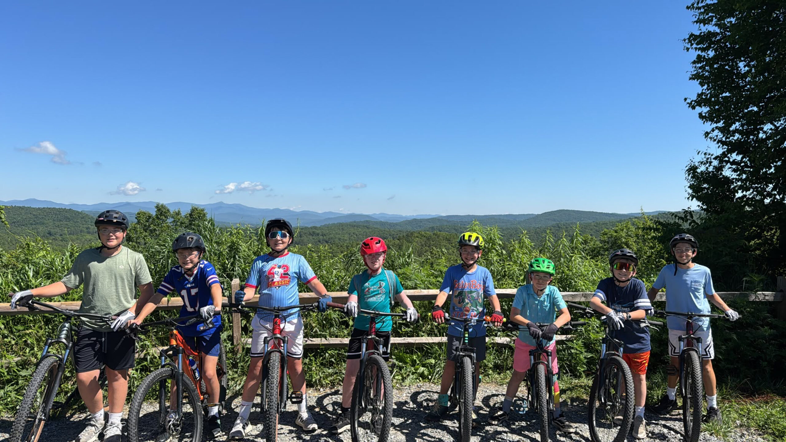 Group of children in cycling gear with helmets standing beside their bikes on a gravel path, with green hills and blue sky in the background.