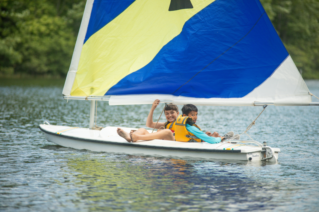 Two kids wearing life jackets on a small sailboat on calm water with a large colorful sail behind them