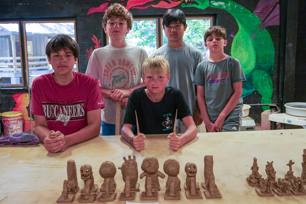 Six kids stand and sit behind a table displaying a row of brown clay figures they molded in a ceramics class.