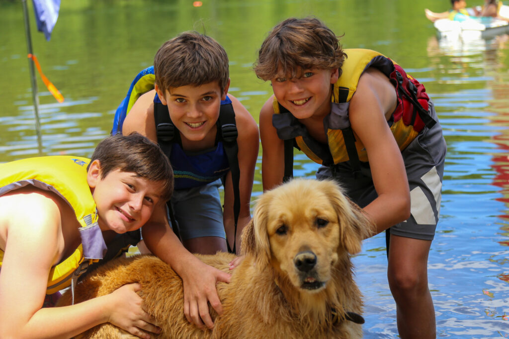 Kids in life jackets gently pet a golden retriever by a lake, smiling at the camera.