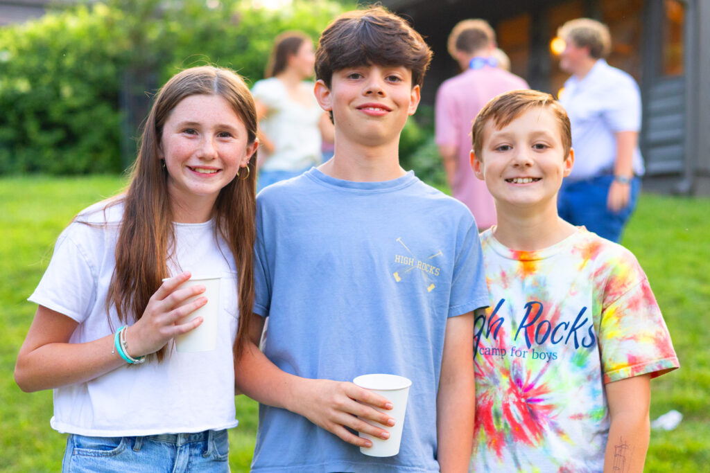 Three smiling kids stand outdoors at a sunny gathering, each holding a white cup (left girl in white, middle boy in blue, right boy in tie-dye).