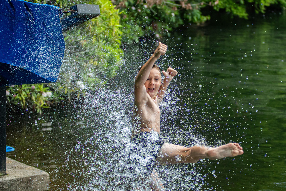 Young boy in swim trunks jumps into a lake from a dock, creating a large splash near green foliage.",
