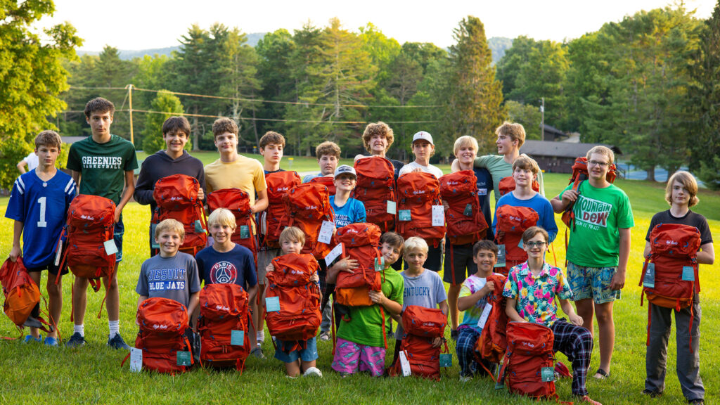 Group of children posing with red backpacks on a grassy field outdoors, smiling at the camera.