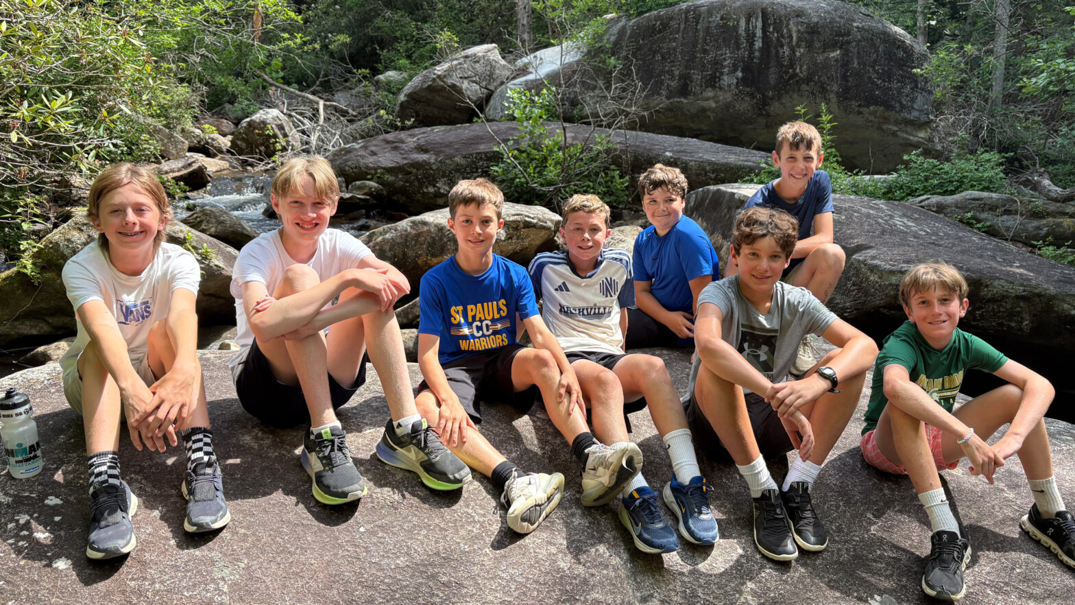 Nine smiling children sit on large rocks beside a forest stream, posing for a group photo.