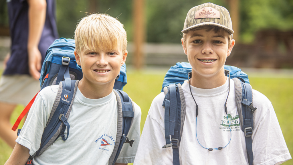 Two smiling boys with backpacks standing outdoors in a field or park.