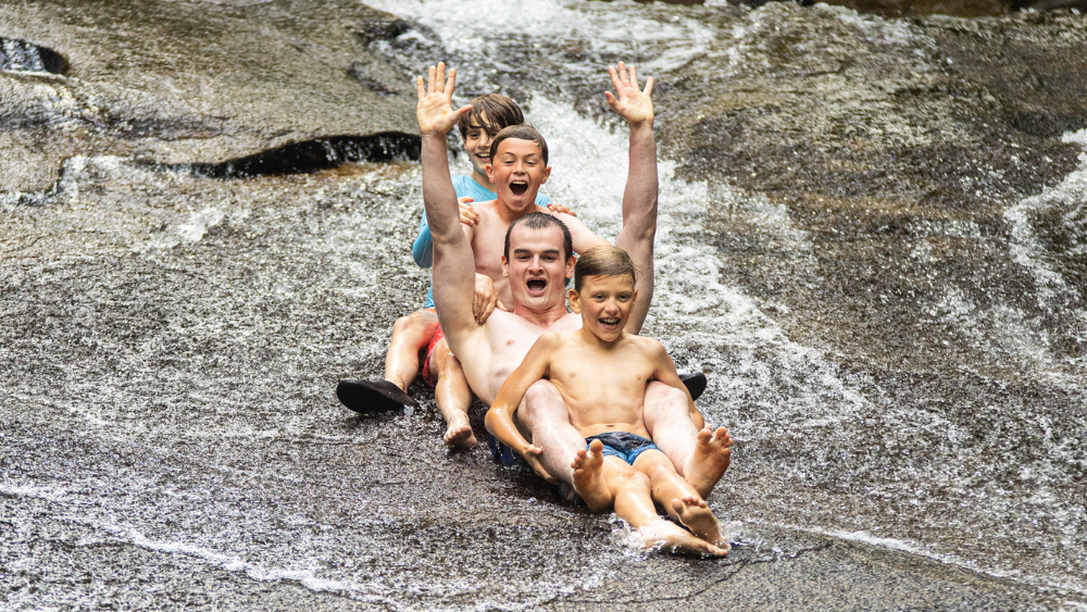 Man and three boys sliding down a wet rock surface, arms raised, grinning playfully on a sunny day.