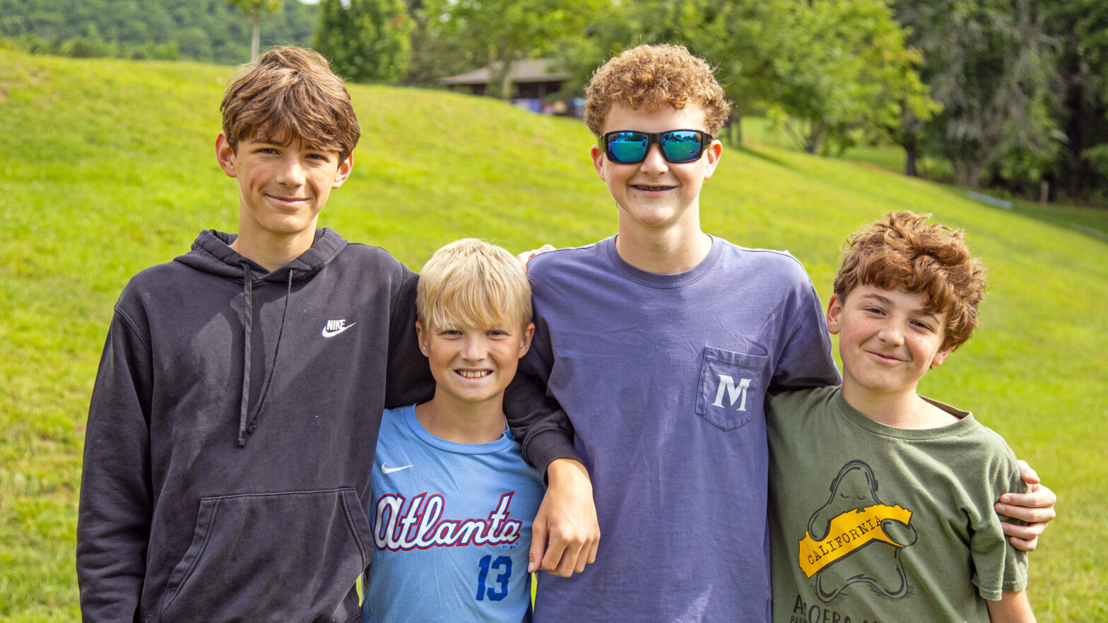 Four smiling boys stand arm-in-arm in a sunlit grassy field, posing for the photo in a park-like setting.