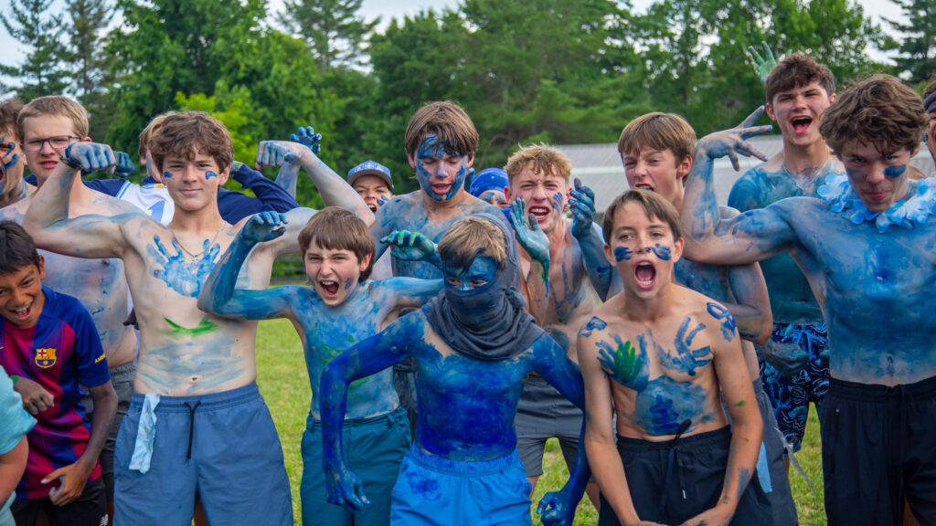 Group of boys outdoors, arms raised and smiling, covered in blue body paint in a grassy field.