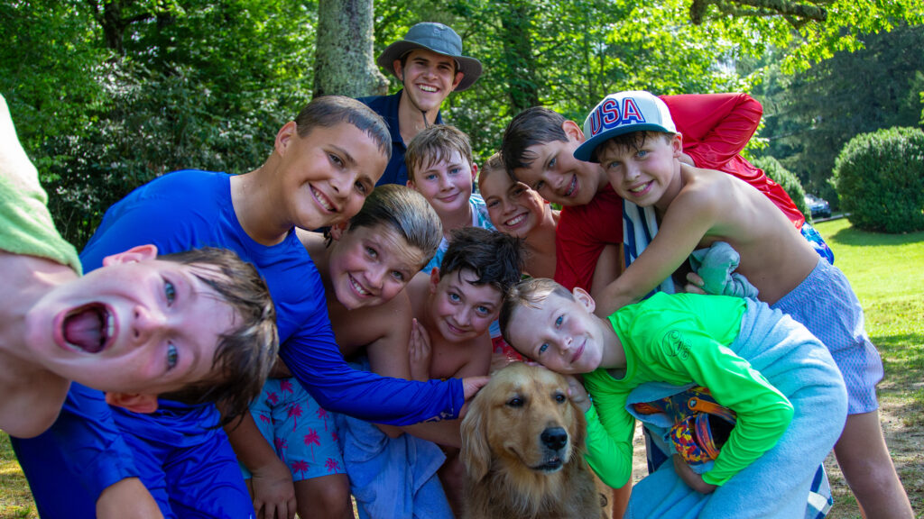 Group of smiling kids in swimsuits and towels posing with a golden retriever outdoors on a sunny day in a park.