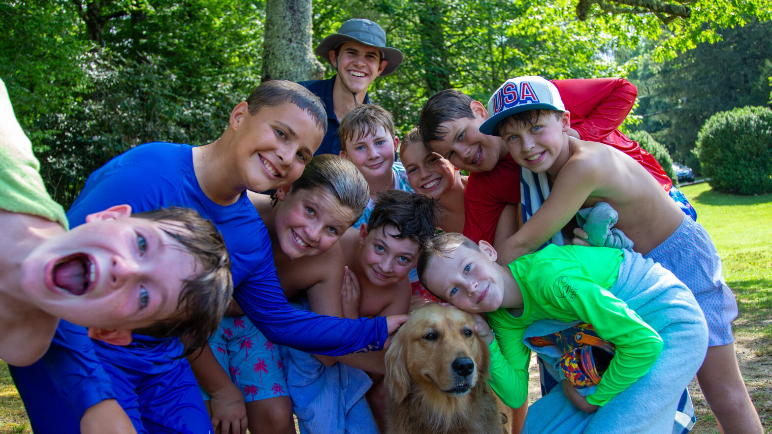 Group of smiling kids in swimsuits and towels posing with a golden retriever outdoors on a sunny day in a park.