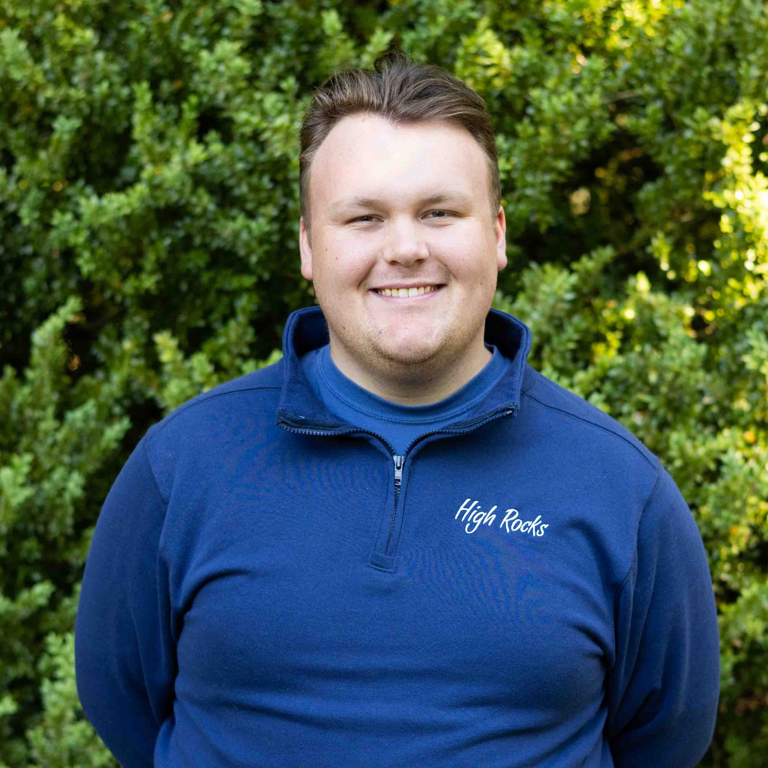 Smiling man in a blue pullover with 'High Rocks' logo, outdoors with green leafy bushes behind him.
