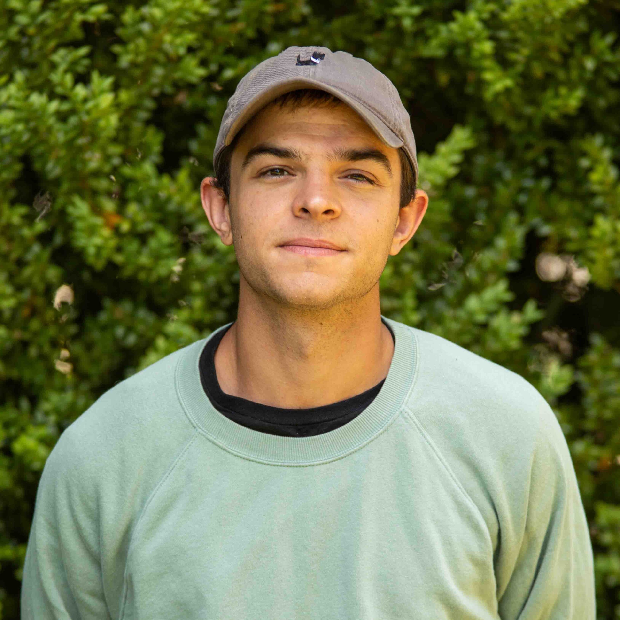 Close-up portrait of a young man wearing a gray cap and mint-green sweatshirt, with green foliage in the background.