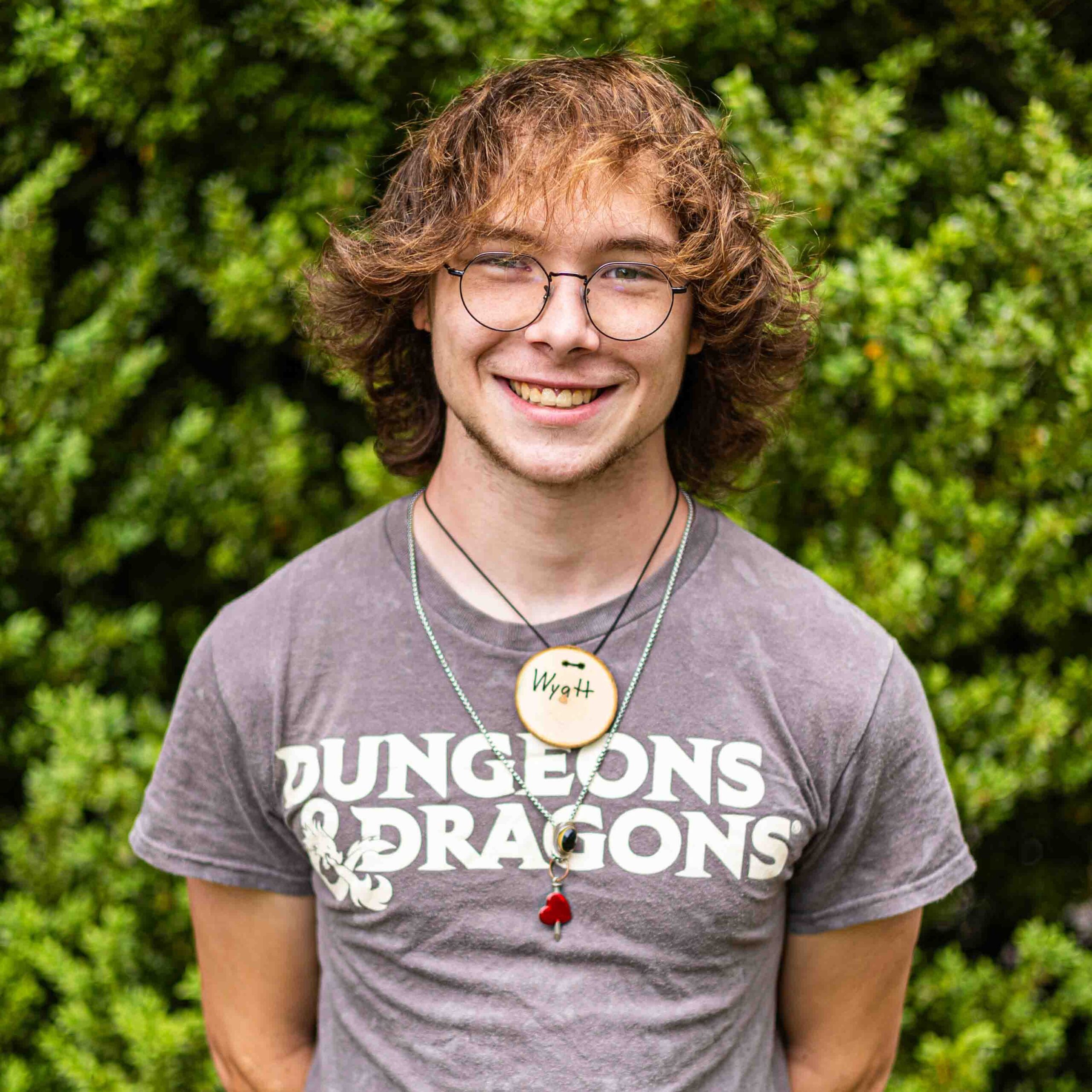 Young person with round glasses and wavy hair smiling outdoors, wearing a gray 'Dungeons & Dragons' shirt.