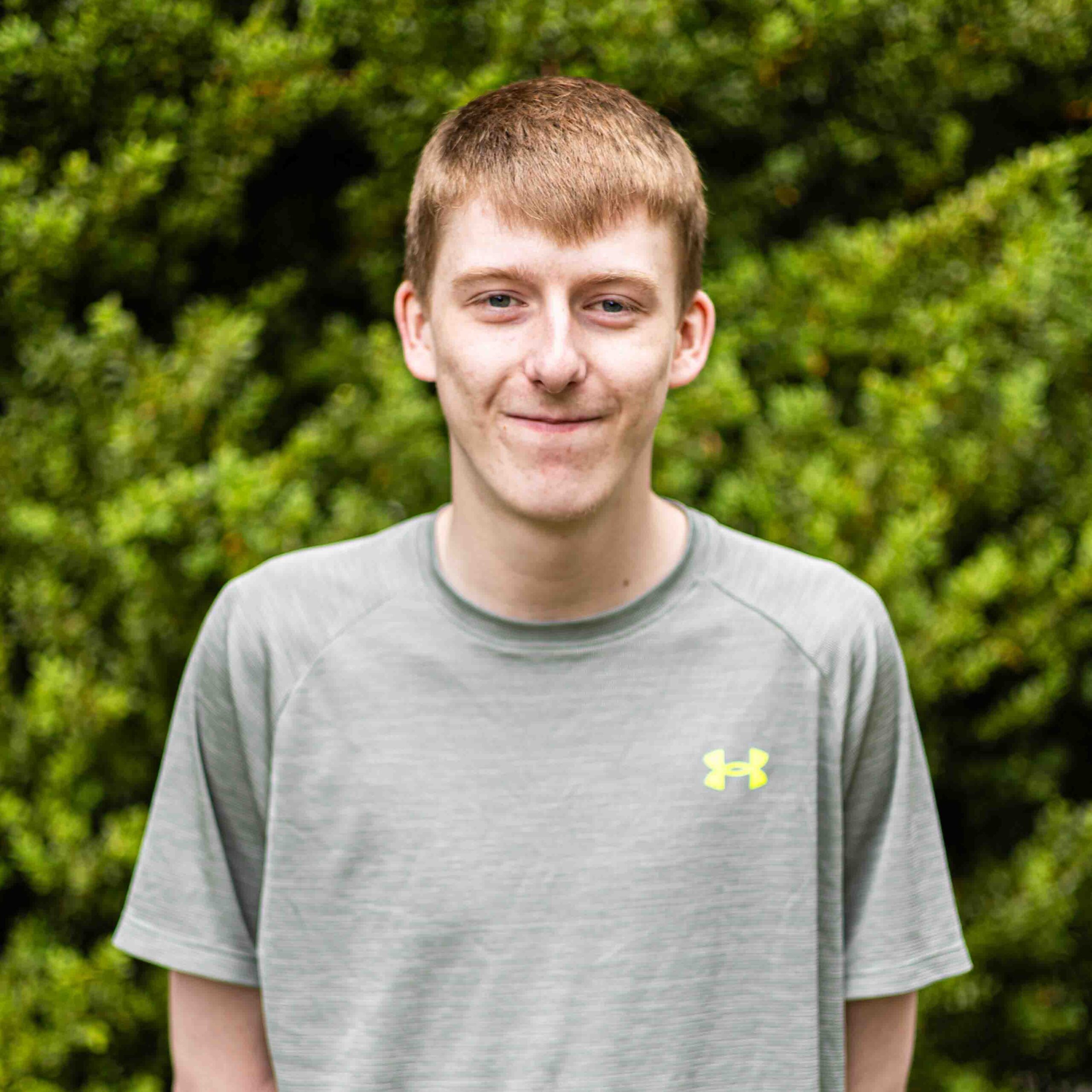 Smiling young man in a light gray athletic shirt standing outdoors with green bushes behind him.