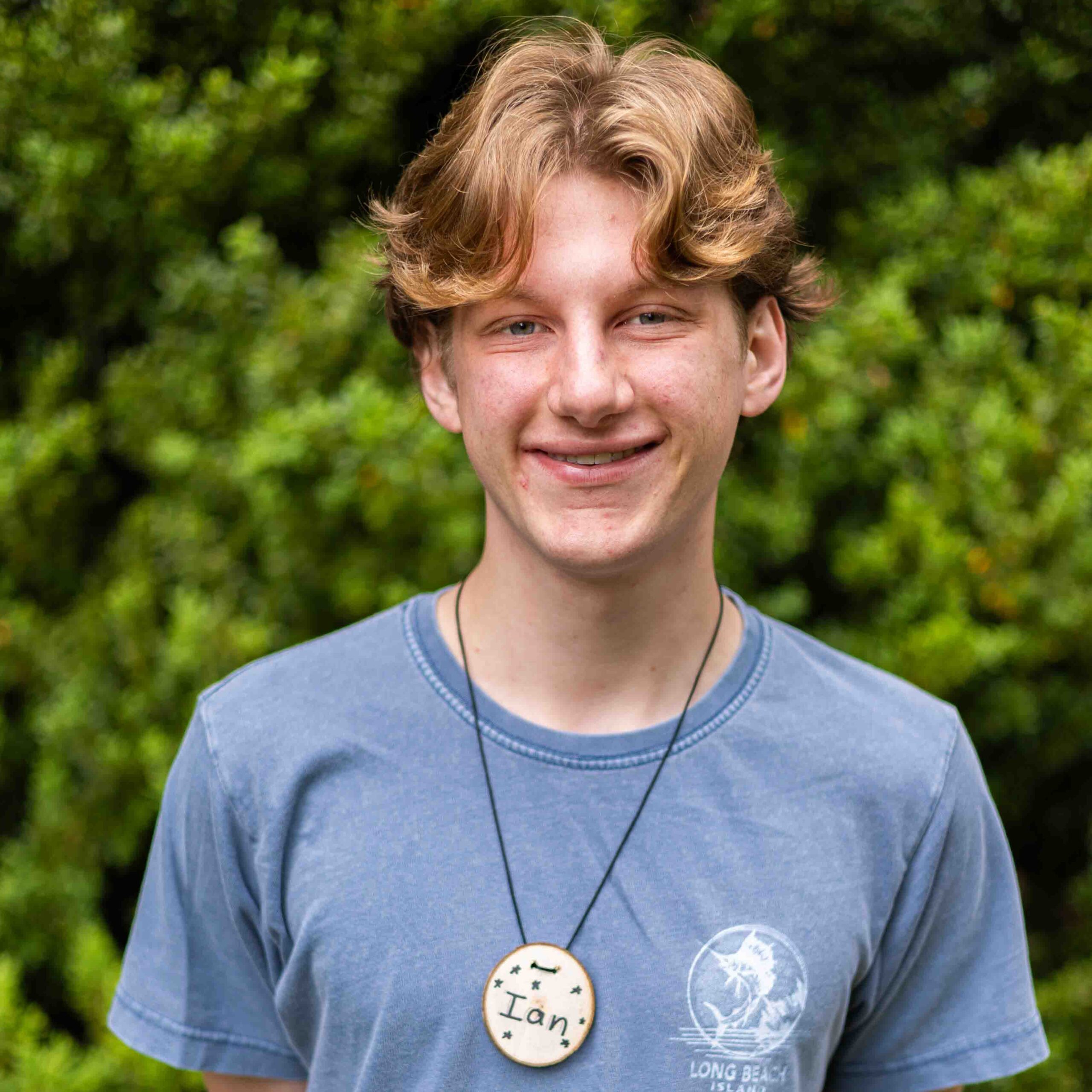 Young man wearing a blue T-shirt with circular badges pinned to chest, standing outdoors with green foliage in background, facing camera.