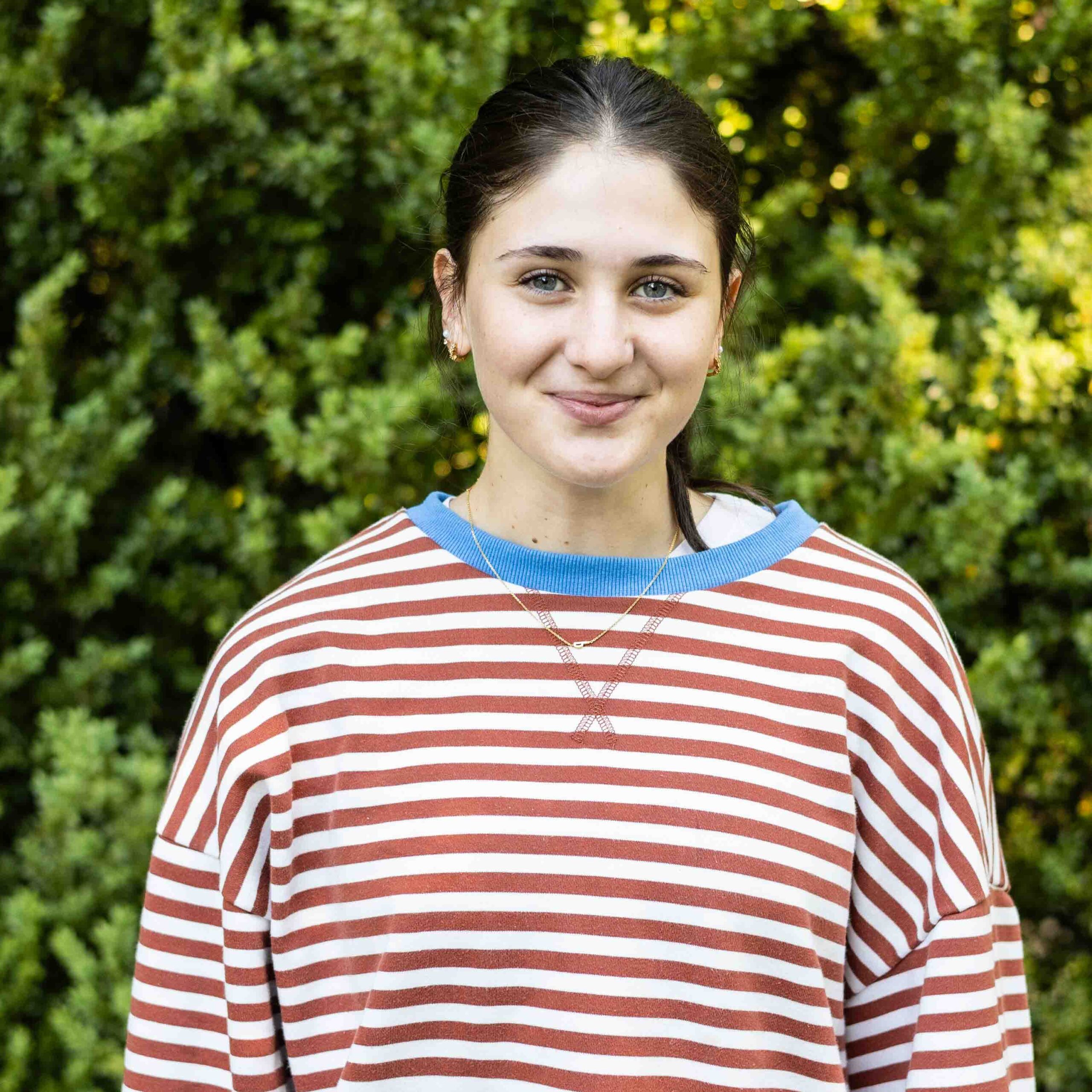 Close-up portrait of a smiling woman outdoors, wearing a red-and-white striped shirt with a blue collar and a necklace.