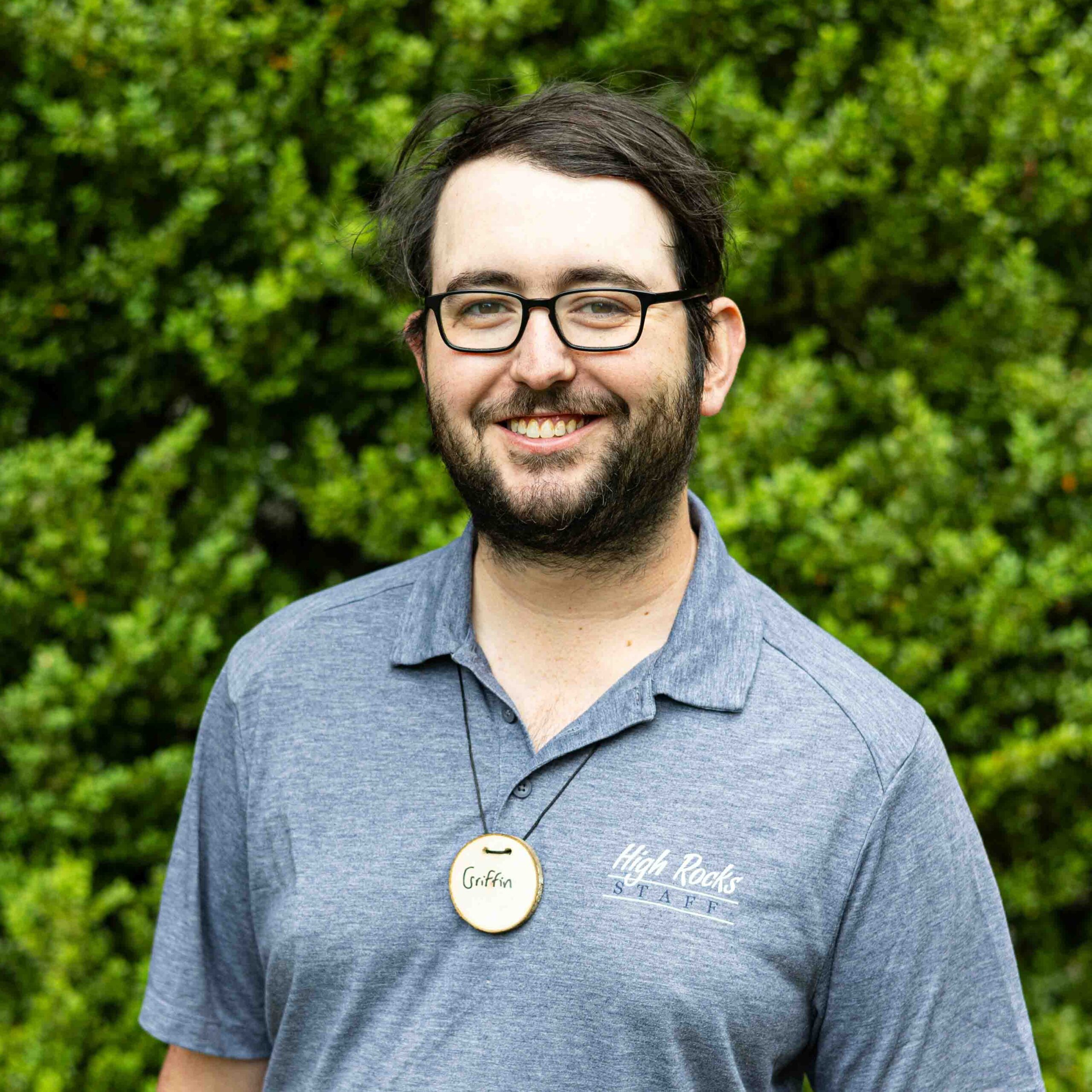 Smiling man with short dark hair and glasses outdoors, wearing a circular name badge that says ‘Griffin’ and a High Rocks staff shirt, with green foliage in the background.