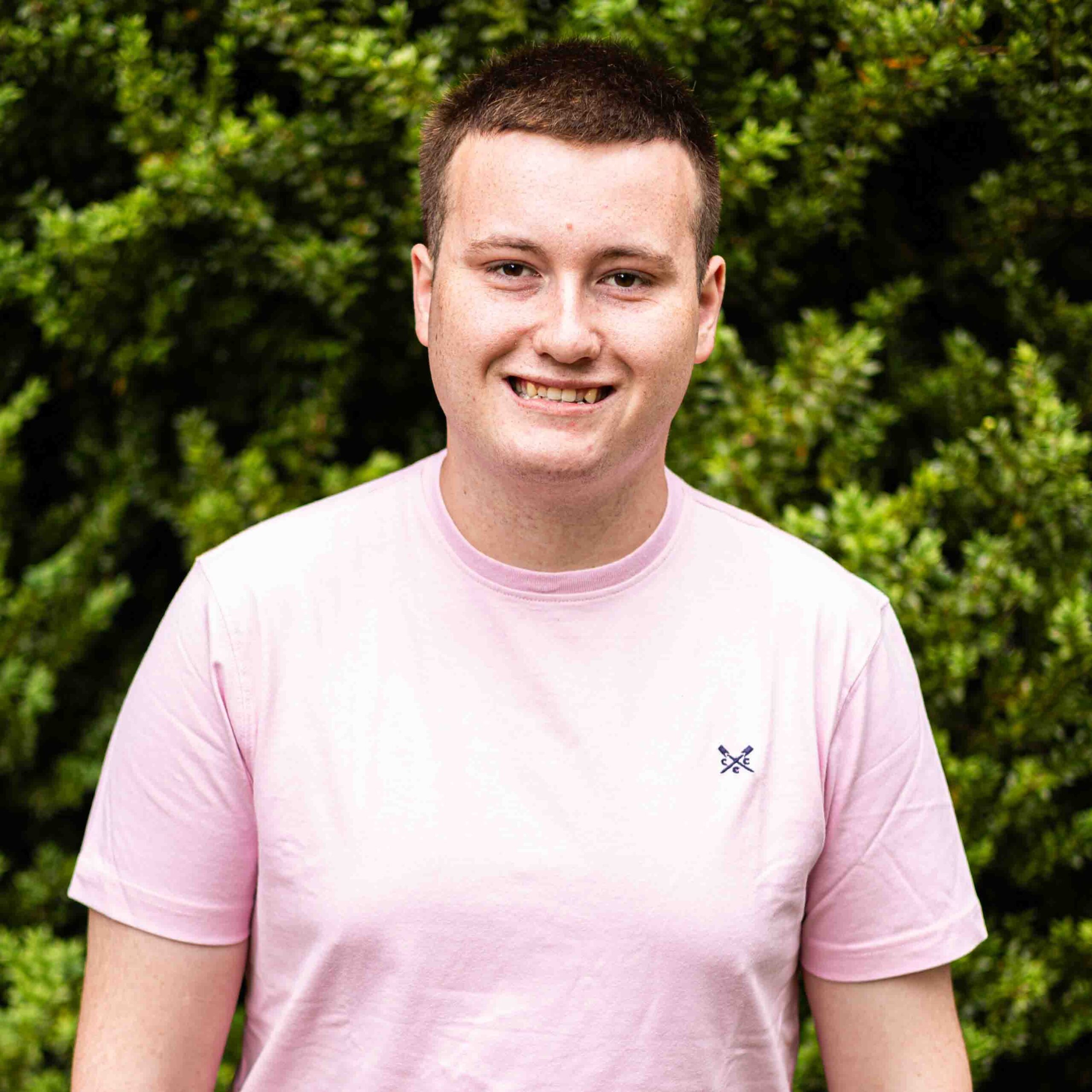Person wearing a pale pink T-shirt standing outdoors with green bushes in the background, smiling slightly.