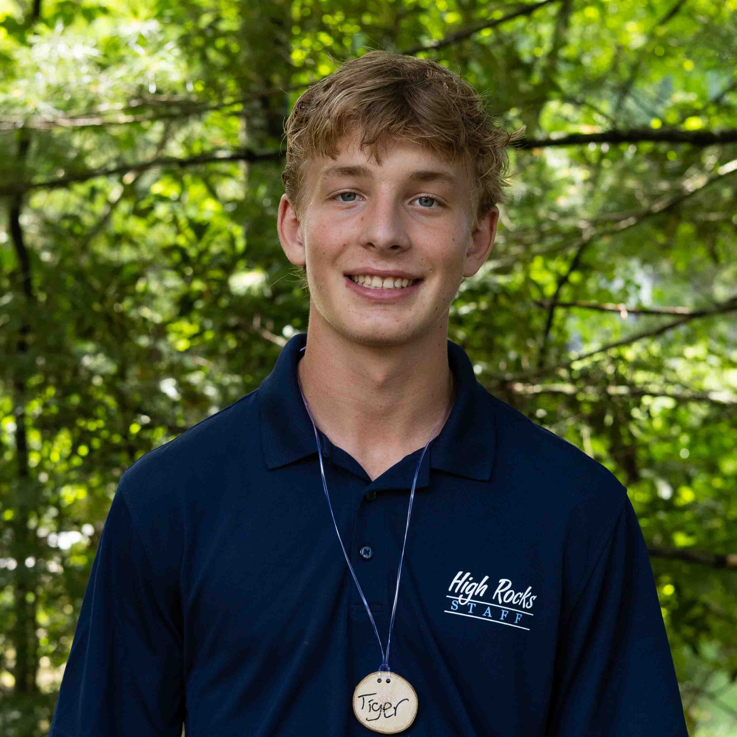 Young man smiling outdoors wearing a navy polo shirt with a circular name tag reading 'Tiger' against a green leafy background.
