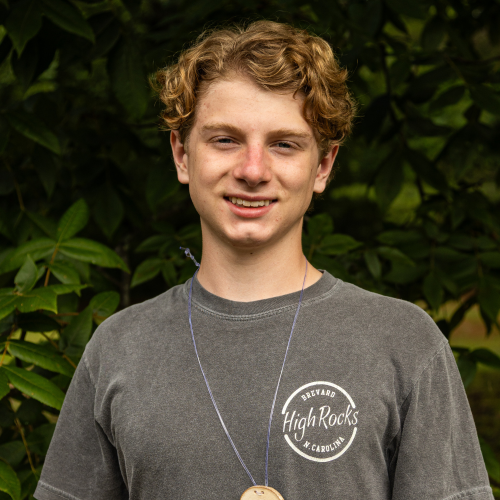 Young man with curly blonde hair wearing a gray High Rocks t‑shirt and a pendant necklace, outdoors with greenery in the background.