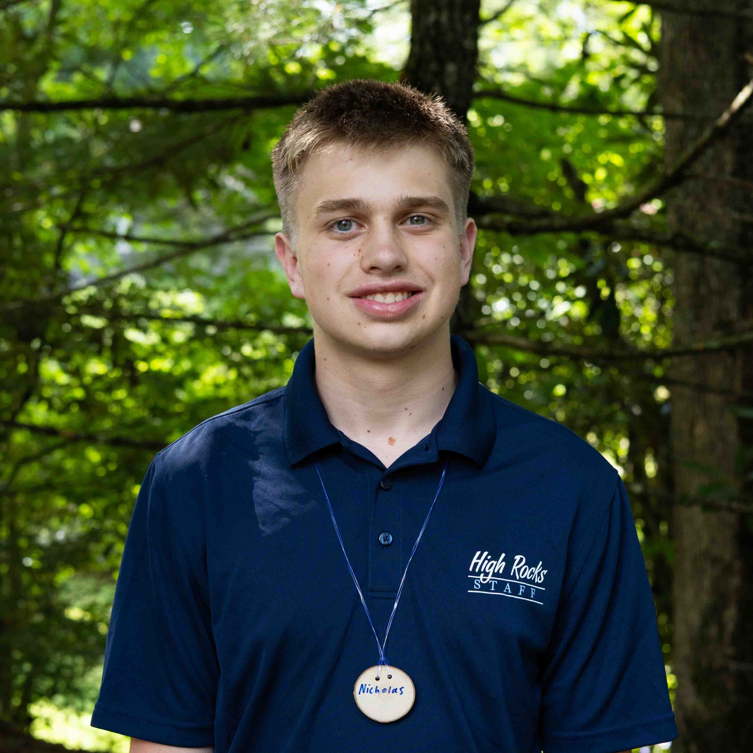 Young man in a navy polo with 'High Rocks Staff' embroidered, wearing a name badge that reads 'Nicholas', outdoors in a leafy setting.