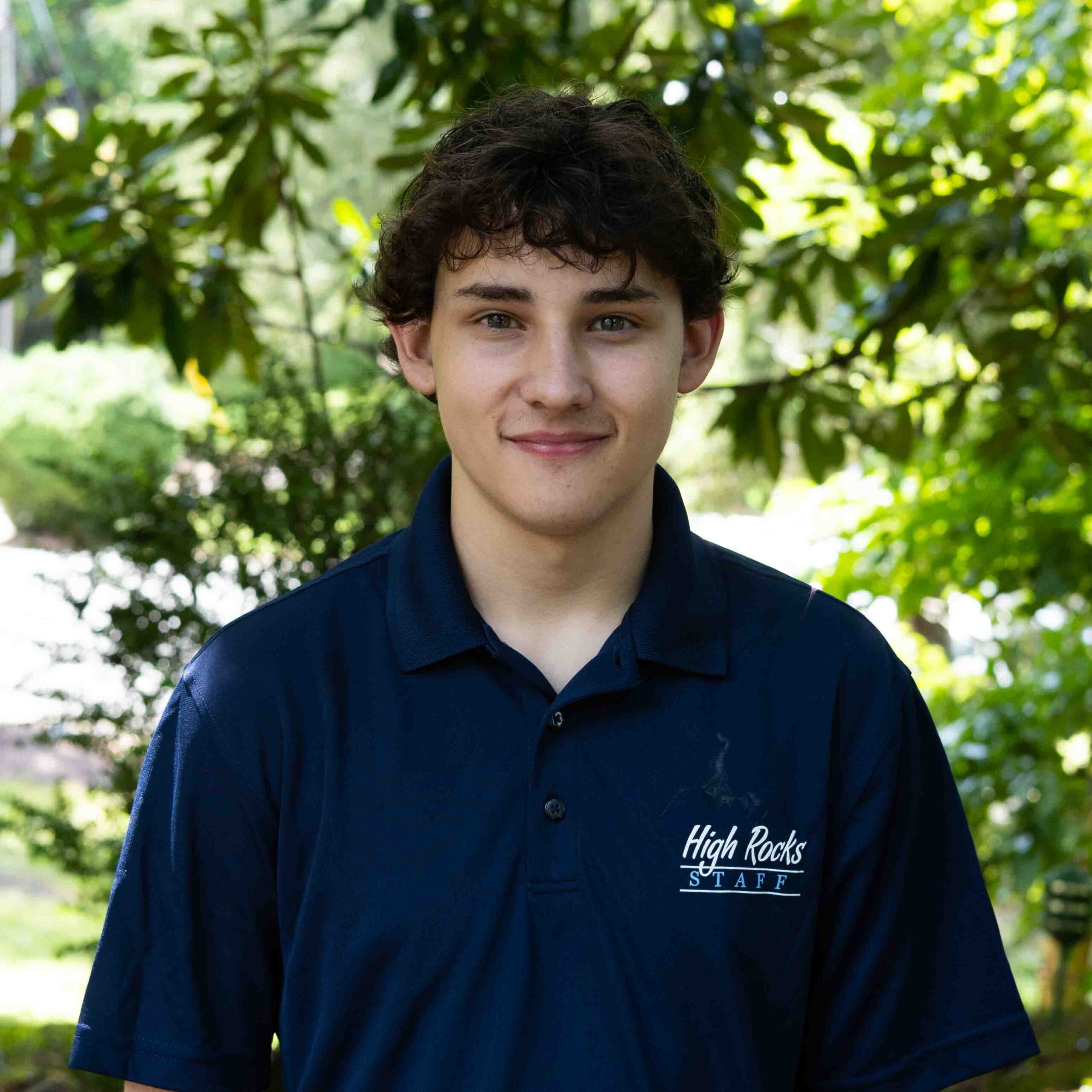 Young man wearing a navy polo with High Rocks Staff logo, outdoors among green trees.
