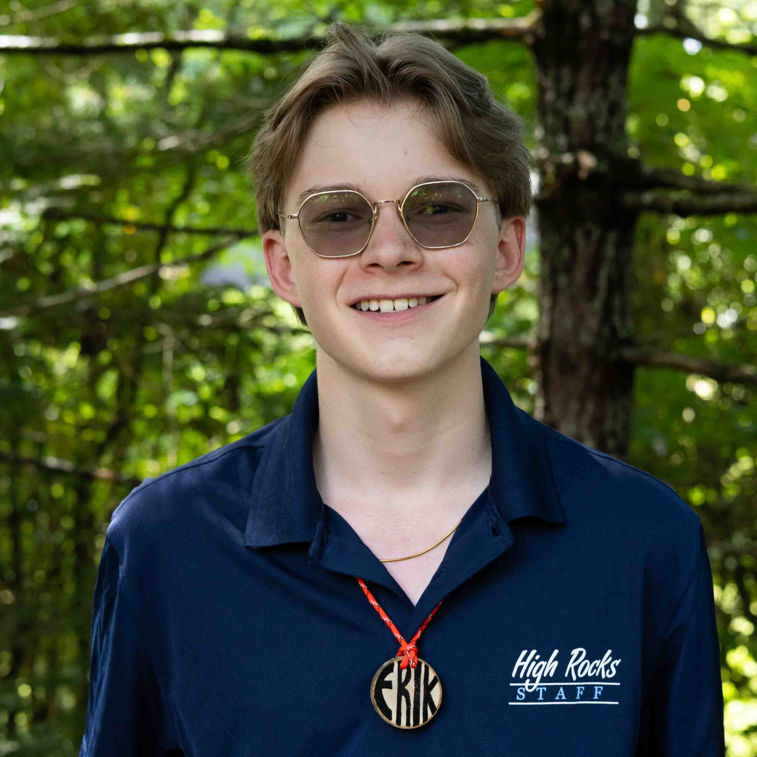 Young person wearing a navy polo with 'High Rocks STAFF' on the chest, outdoor wooded background, wearing round glasses and a red lanyard with a badge.