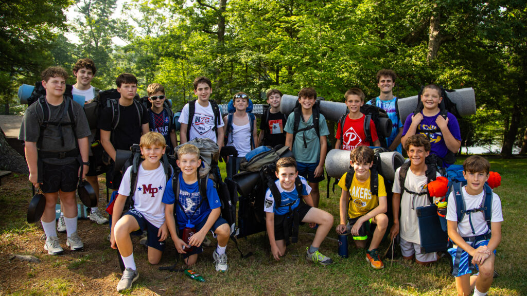 Group of smiling children wearing backpacks and camping gear, posing for a photo outdoors among trees in a park.