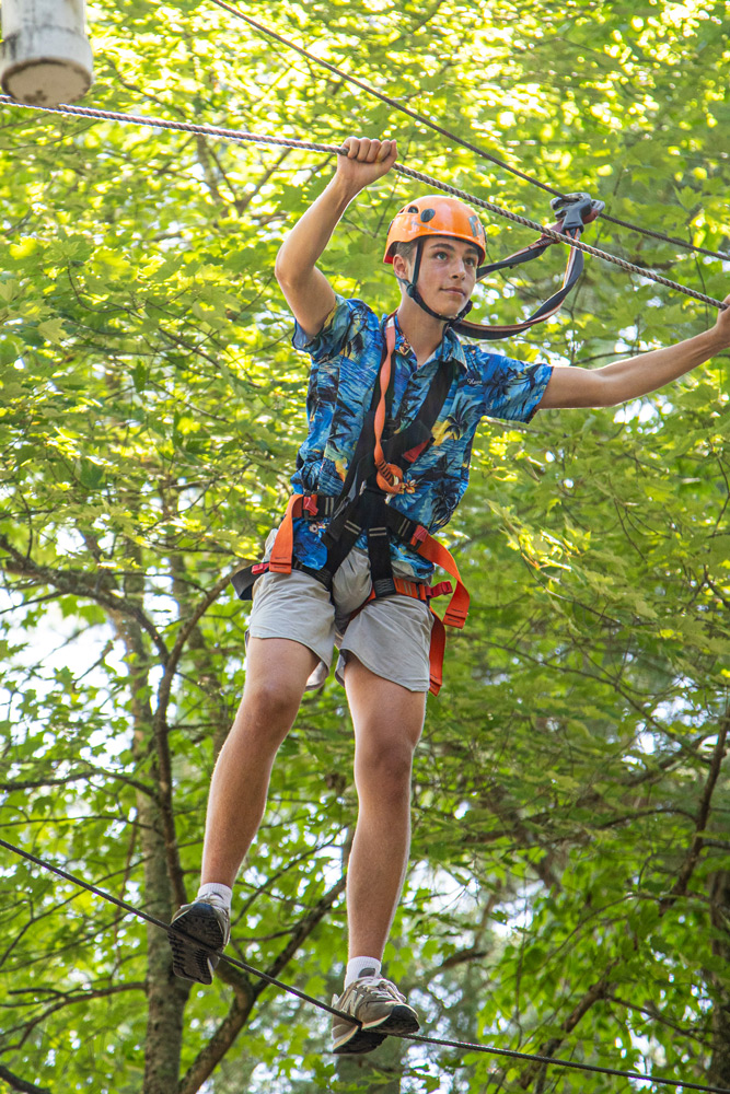 Person wearing an orange helmet and safety harness traversing a high ropes course among green trees, gripping overhead ropes.