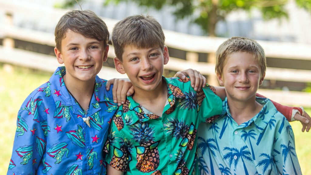 Three boys in colorful tropical shirts with arms around each other, smiling outdoors.
