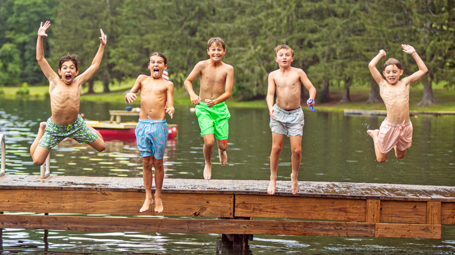 Five shirtless boys leap off a wooden dock into a lake, laughing in colorful swim trunks with trees behind them.