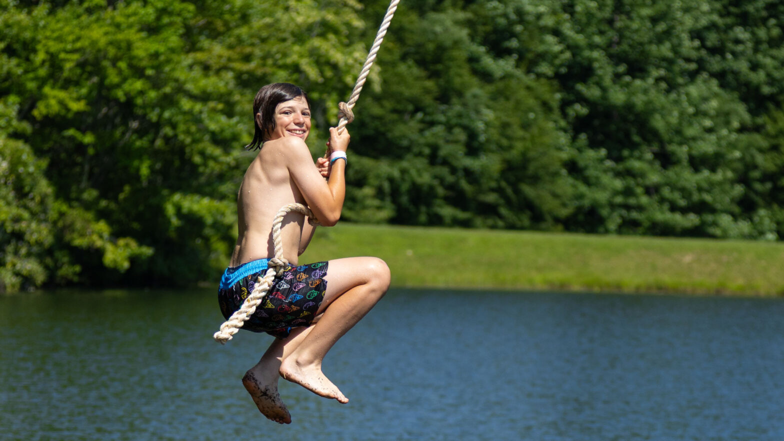 Person swinging on a rope over a lake, smiling with green trees in the background.