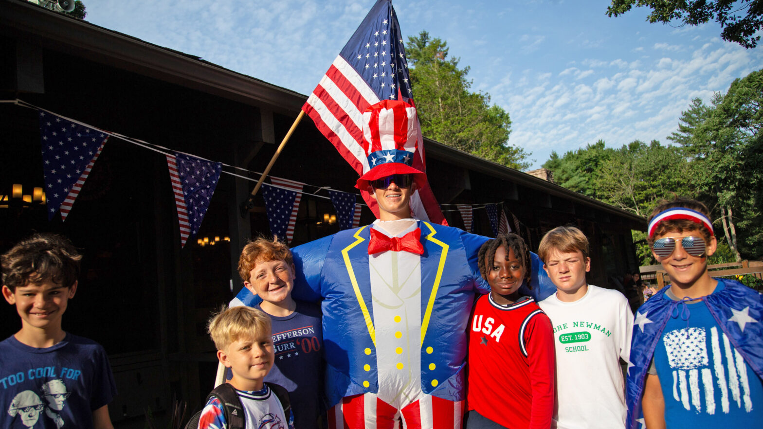 Group of smiling kids posing with a person in a giant Uncle Sam costume wearing an American flag hat at a patriotic outdoor event.