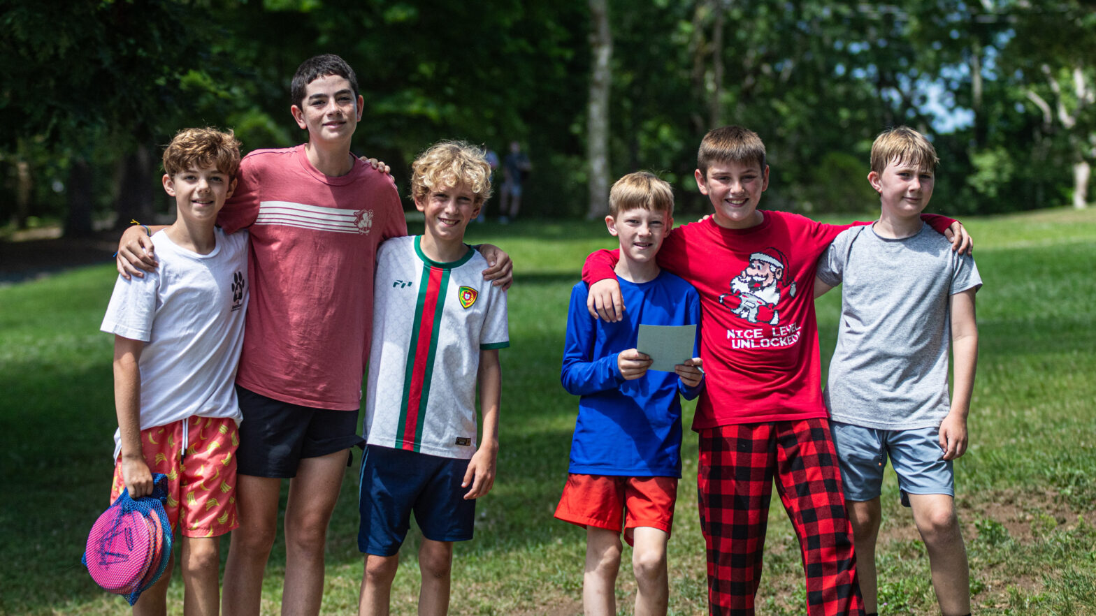 Six boys stand together in a grassy park with trees, arms around each other's shoulders, smiling at the camera.