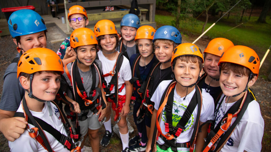 Group of kids in orange and blue helmets wearing climbing harnesses, posing for a photo outdoors at a camp.