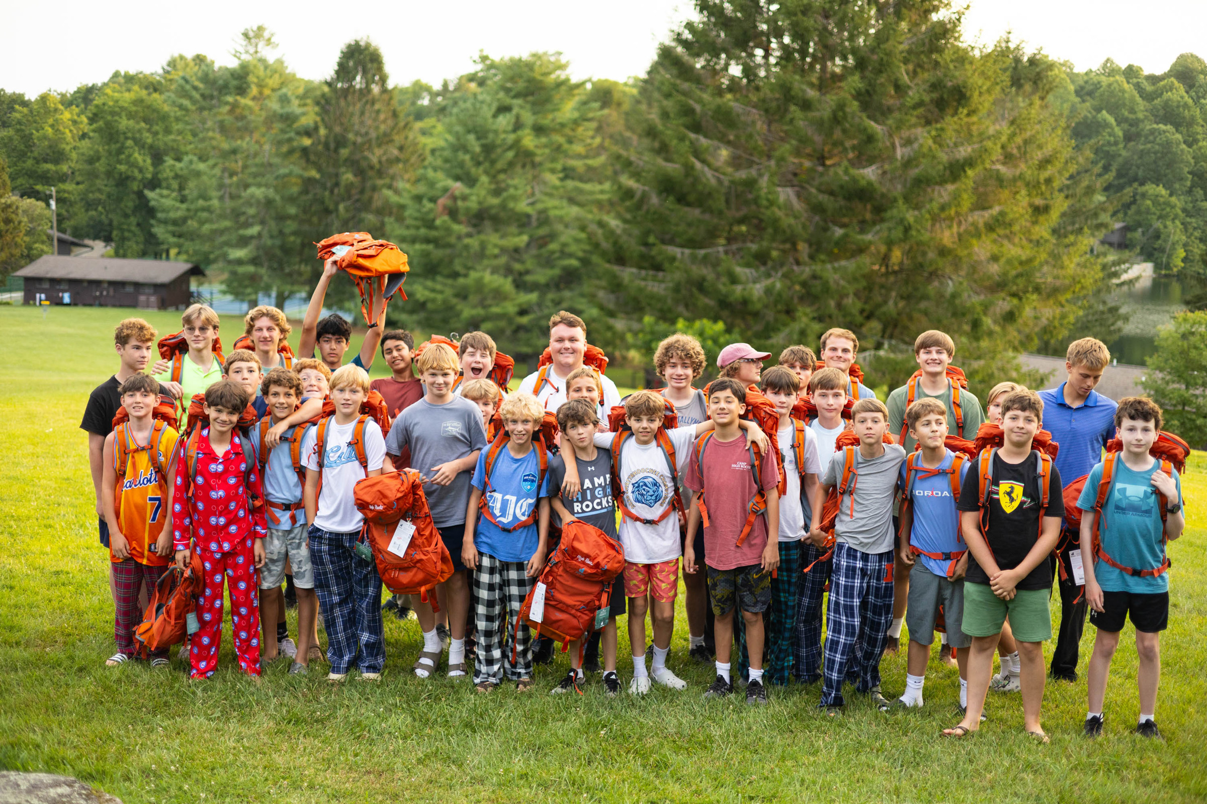 Young campers in orange backpacks stand together on a grassy field with trees in the background, smiling for a group photo.