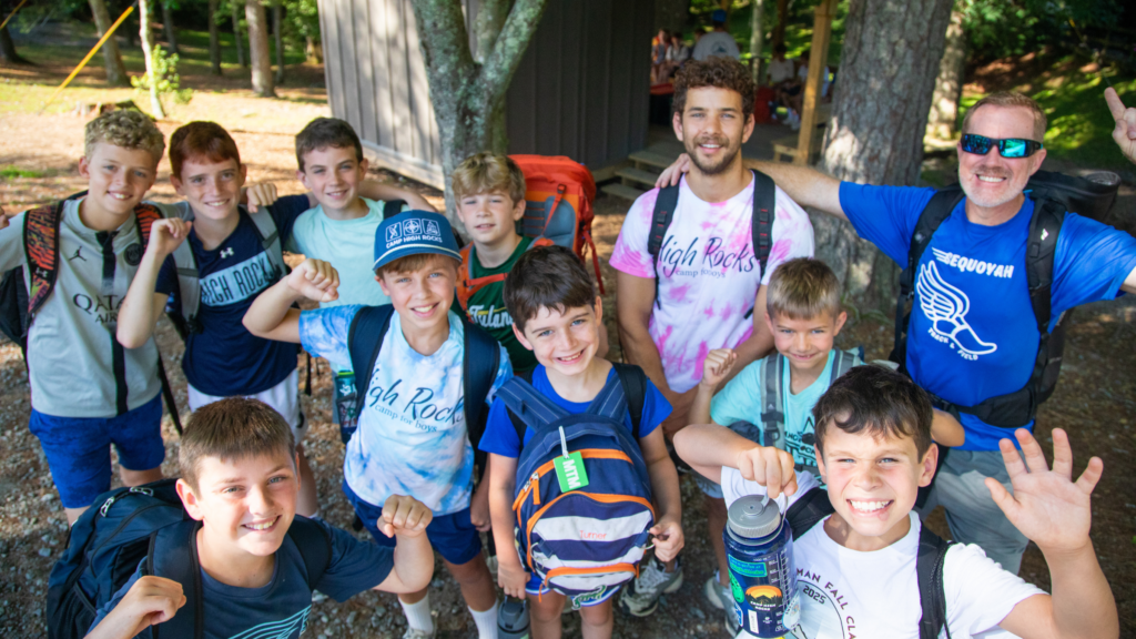 Group of smiling boys with backpacks posing for a selfie outdoors, a camp counselor stands with them under trees near a wooden cabin.