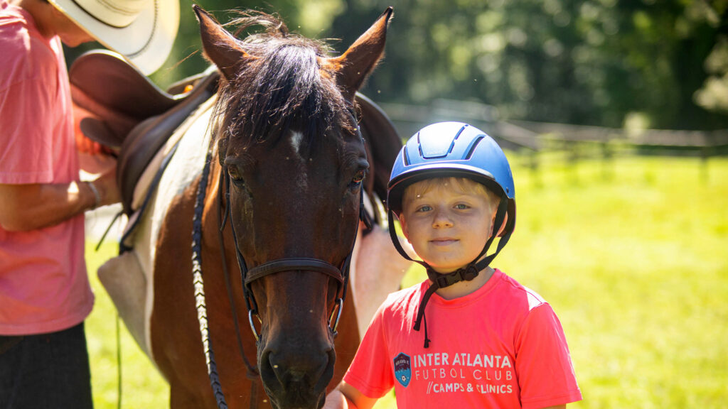 Boy in a pink shirt and blue riding helmet standing beside a dark horse in a sunny field.