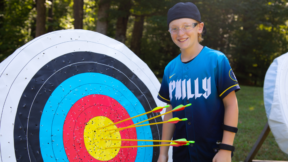 Young archer in a blue jersey and glasses stands beside a colorful archery target with arrows in the bullseye.