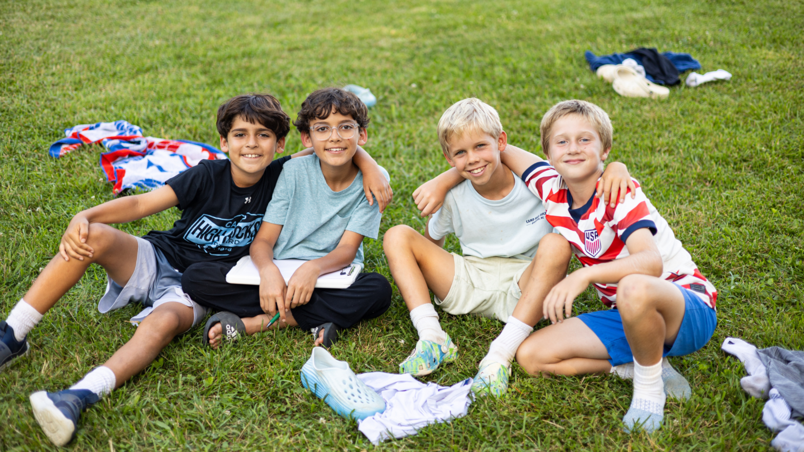 Four smiling boys sit on a grassy field with arms around each other, enjoying a sunny day at the park.