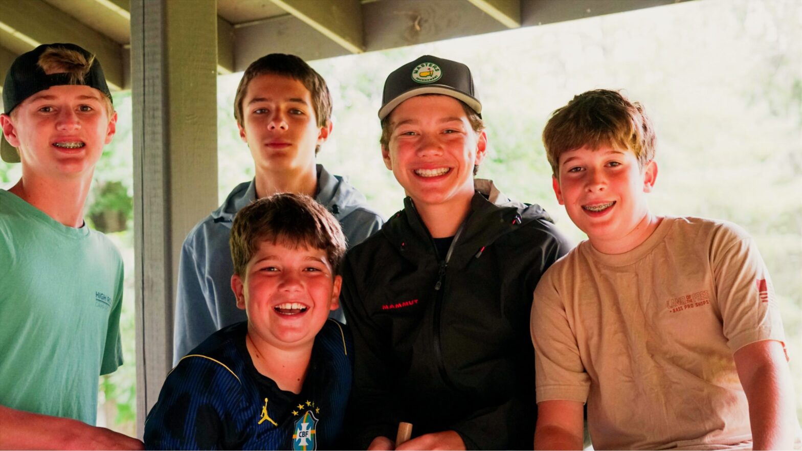 Five teenage boys posing together under a wooden shelter, smiling at the camera. 5 people in casual shirts and jackets activity outdoors under a pavilion.