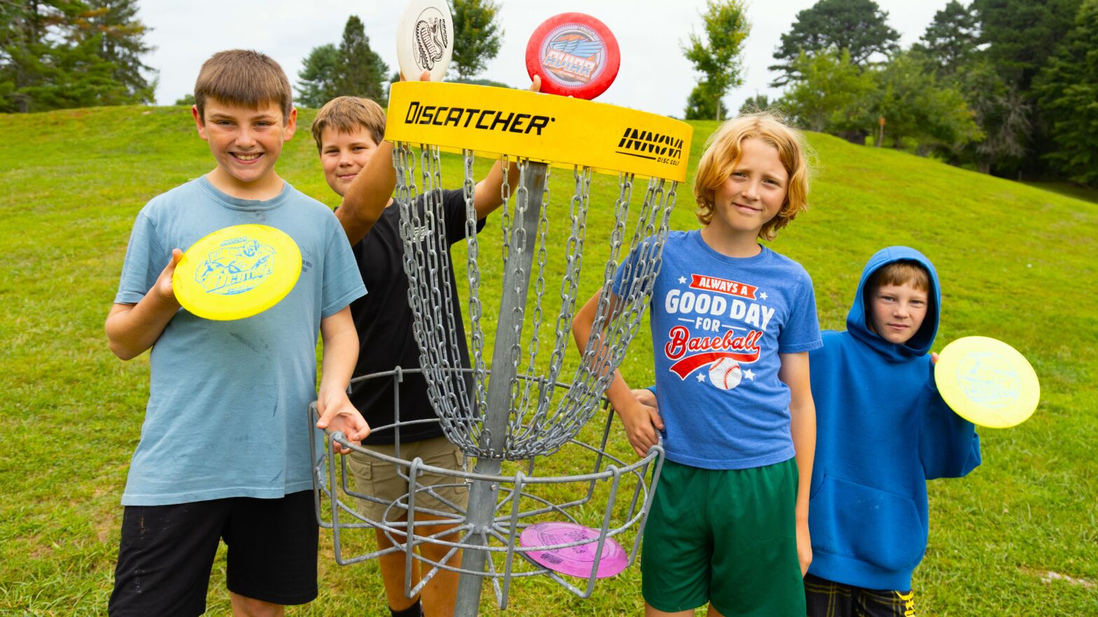 Four children stand around a disc golf basket outdoors, each holding colorful discs and smiling at the camera.
