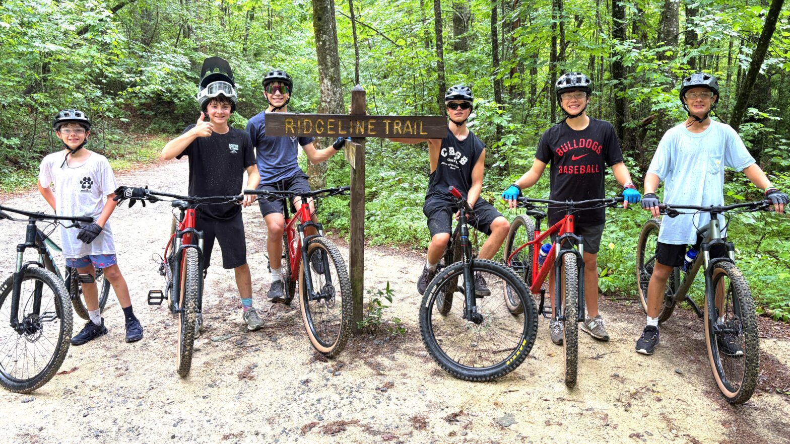 Seven children in helmets with mountain bikes standing on a dirt forest trail beside a wooden 'Ridgeline Trail' sign, smiling at the camera.