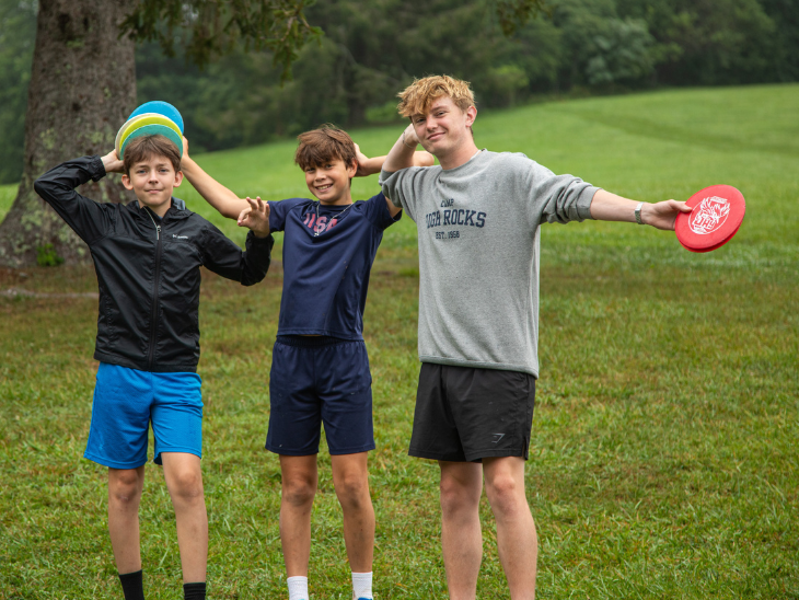 Three boys in a park pose with arms around each other, holding colorful frisbees and smiling at the camera.