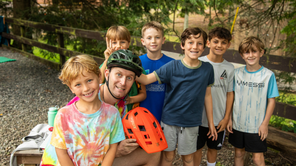 Group of smiling kids and an adult in a bike helmet posing outdoors by a wooden fence and trees.