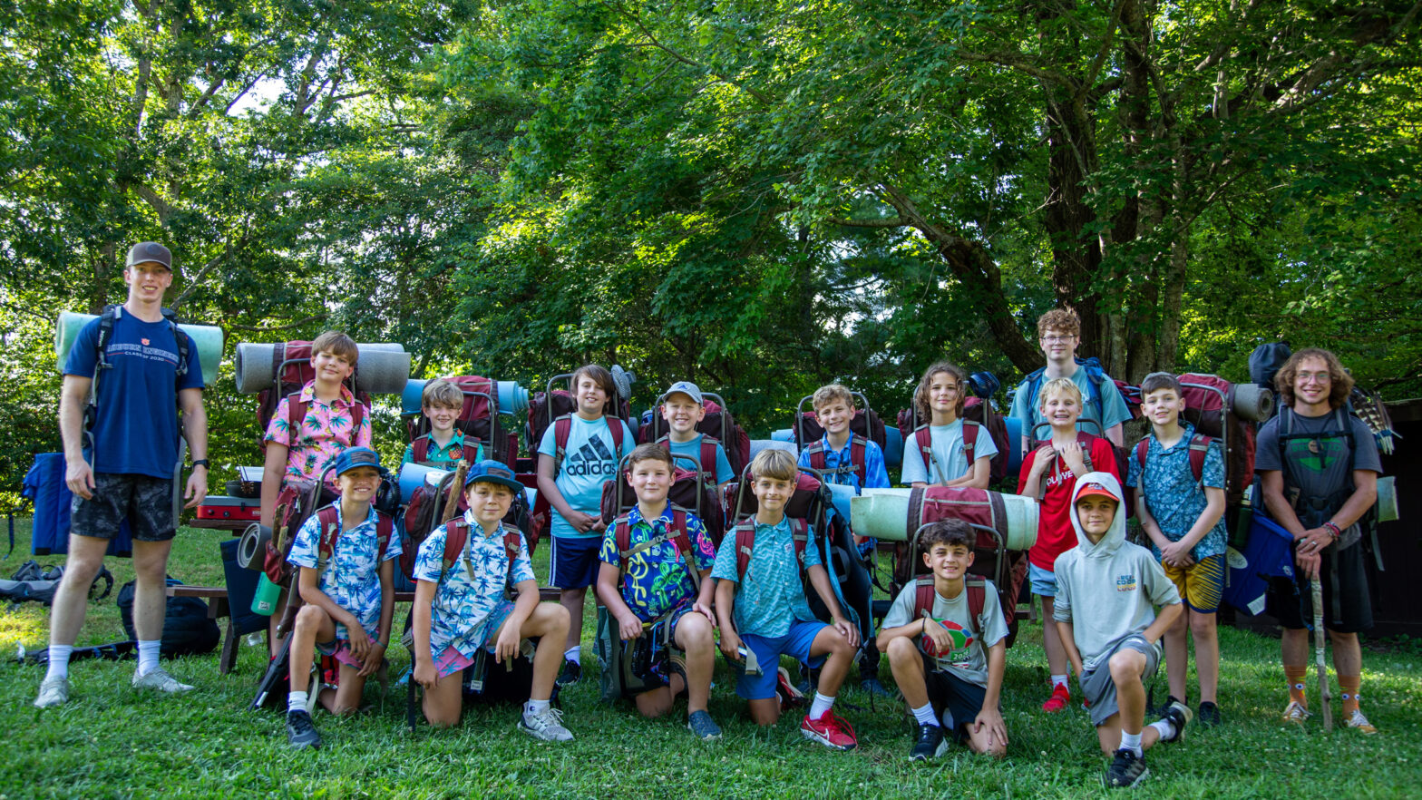 Group of smiling kids and two adults with large backpacks and camping gear in a sunny park with trees in the background.