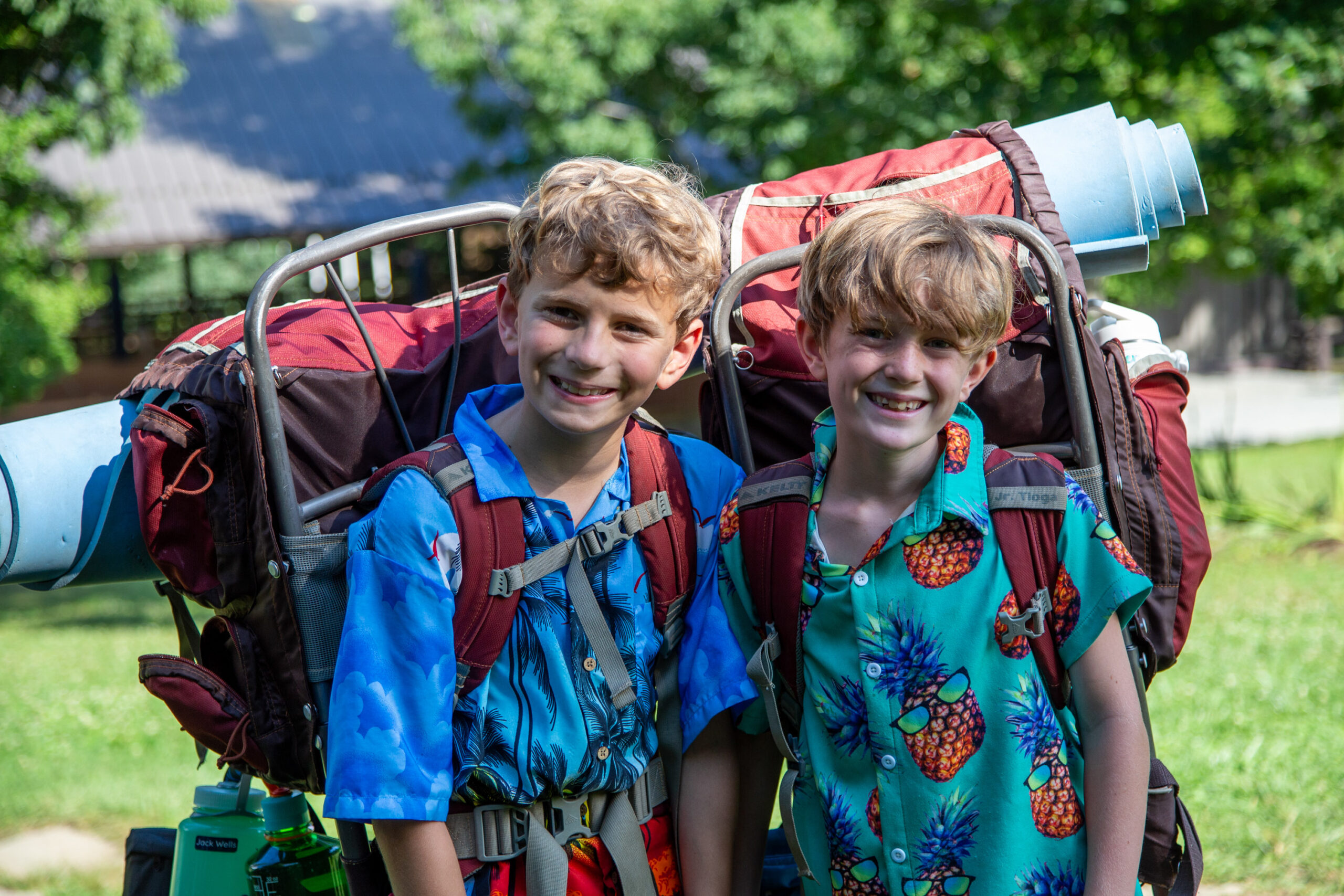 Two smiling boys in bright Hawaiian shirts with backpacks and harnesses stand in a grassy field beside a blue paraglider setup.